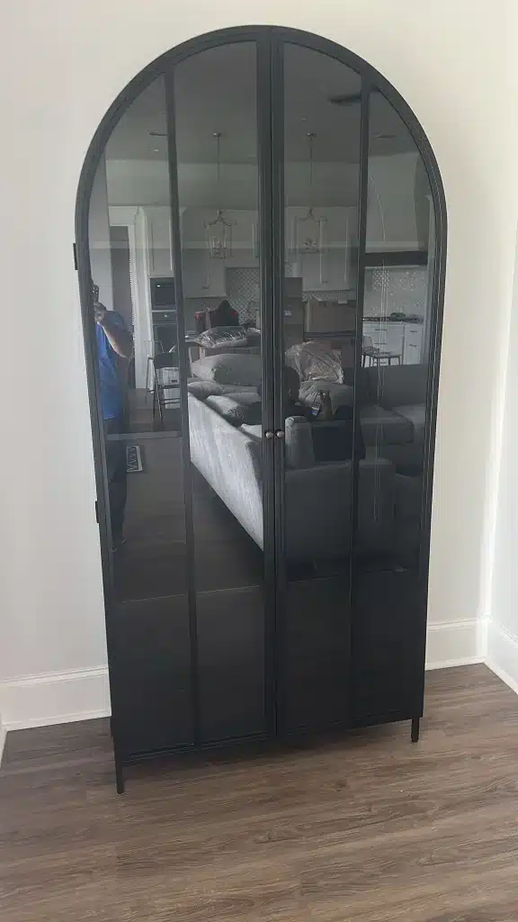 Black arched glass cabinet with metal frame standing against a white wall on wood flooring.