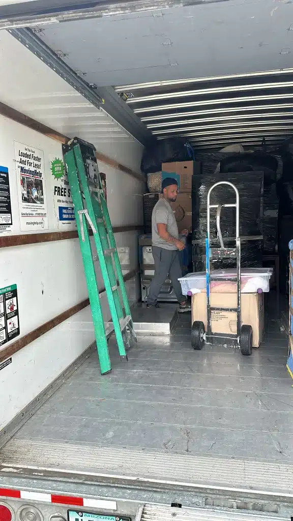 Mover organizing boxes and furniture inside a moving truck with a dolly and green ladder nearby.