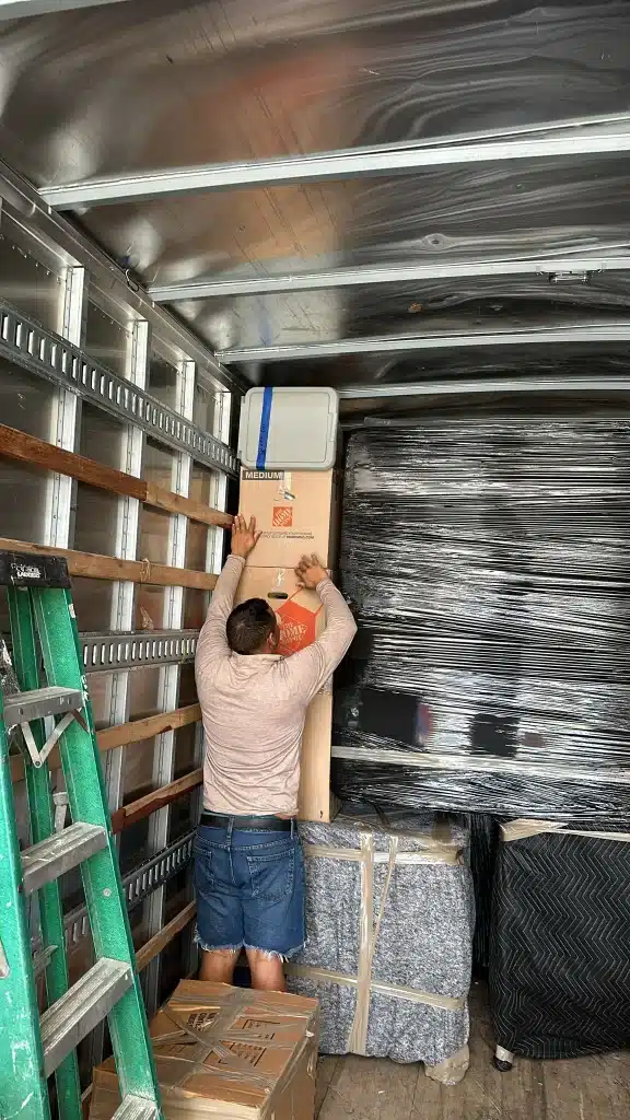 Mover stacking cardboard boxes and wrapped furniture inside a moving truck for transport.