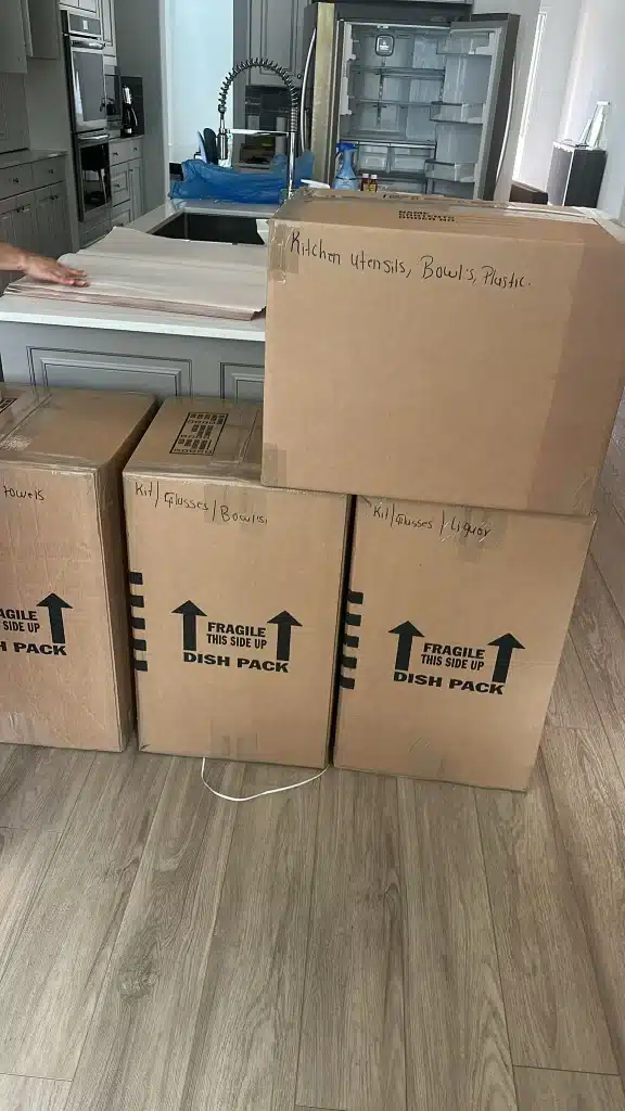 Labeled moving boxes stacked in a kitchen near an island and open refrigerator, ready for unpacking.