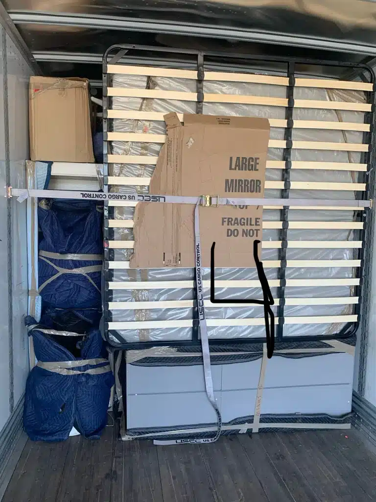 Moving truck loaded with packed furniture, bed frame, boxes, and a labeled cardboard marked “large mirror fragile.”