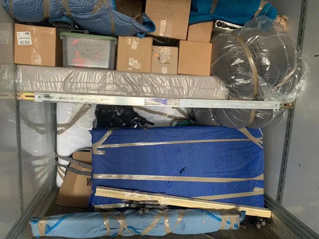 Loaded moving truck with packed boxes, wrapped furniture, mattresses, and blue moving blankets secured with tape.